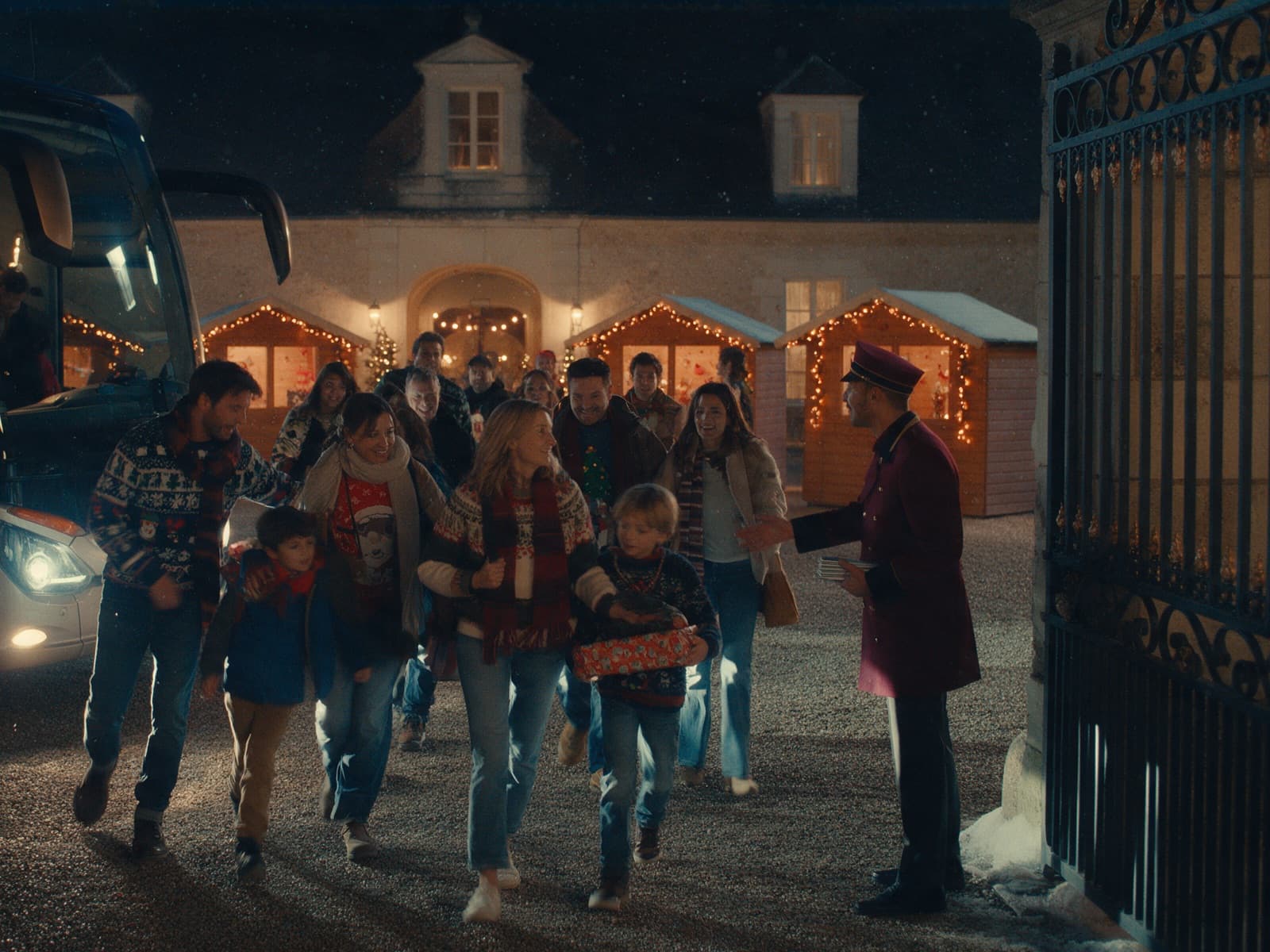 Un groom en tenue bordeaux accueille au portail une bande de collègues et d’enfants en pulls de Noël, chalets éclairés derrière eux.