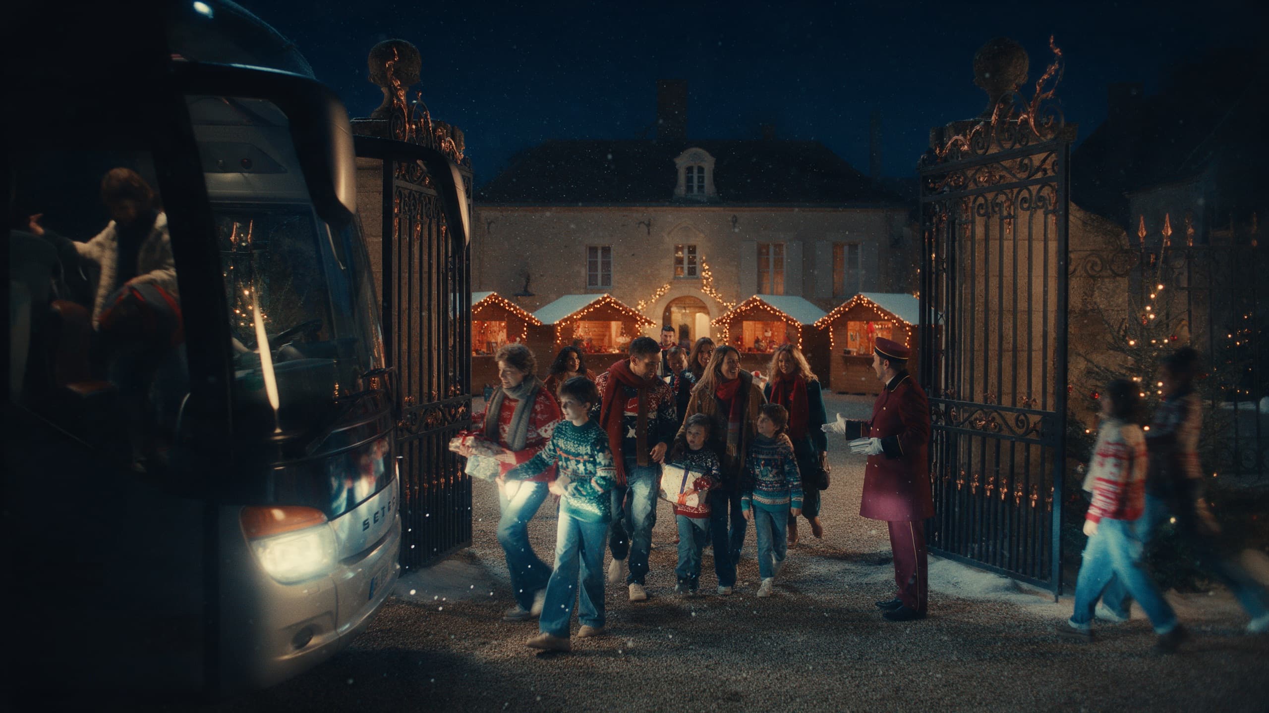 Une bande de collègues et d’enfants en pulls de Noël franchit de nuit le portail du Domaine les 12 Ponts, descendue d’un bus et accueillie par un groom en tenue bordeaux devant quatre chalets éclairés.